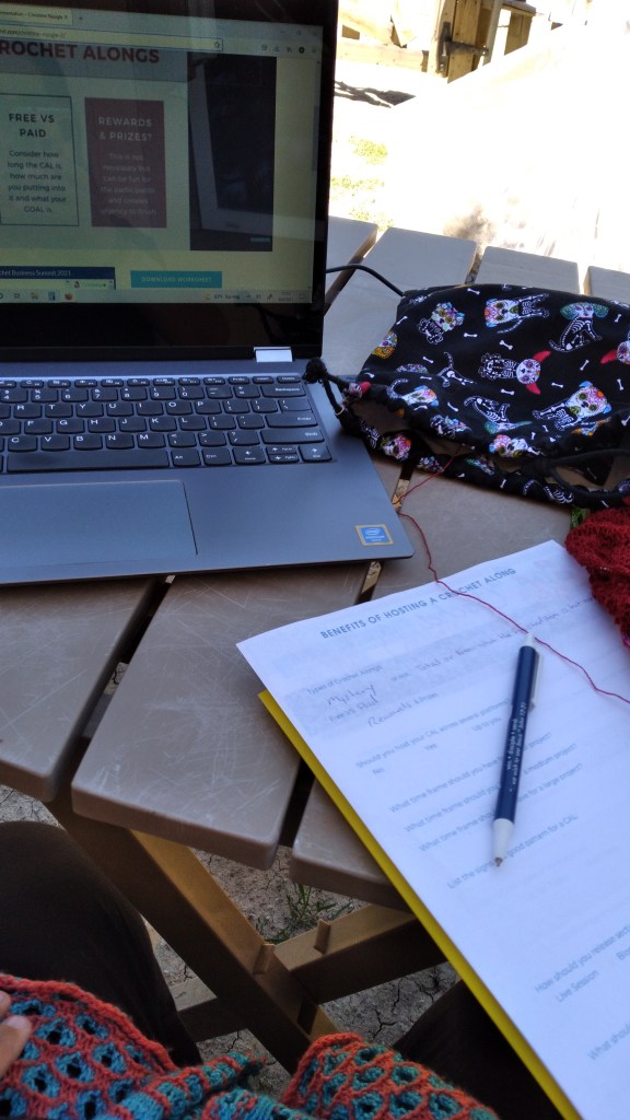 Sara's computer is open on a table outside.  She has her notes for this session of the Crochet Business Summit and her red lace knitting project is off to the right side.