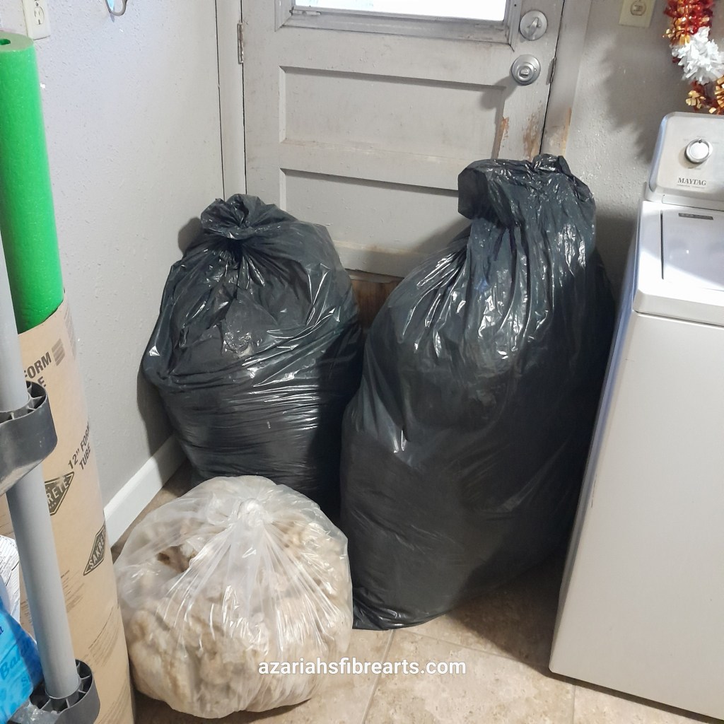 Two large black trash bags and one smaller clear bag are full and tied up sitting on the floor next to a washing machine.