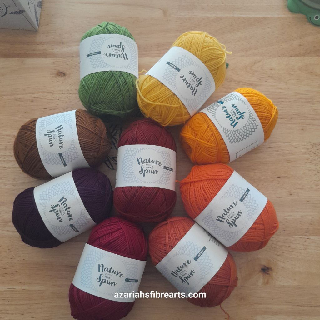 Nine balls of yarn are time are sitting on a brown background. The center ball is a dark red. The surrounding eight balls are all fall colors, in line around the center ball from brown to green, including purple, red, oranges and yellows.