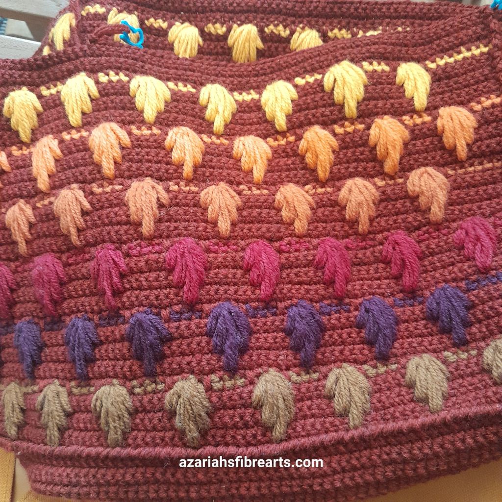 Close up of a crocheted piece. The tight background fabric has stitches of leaves in five different colors from brown to yellow from bottom to top.