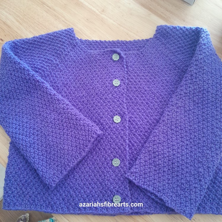A purple knit sweater, with the sliver buttons buttoned and the sleeves slightly folded sit on a brown background.