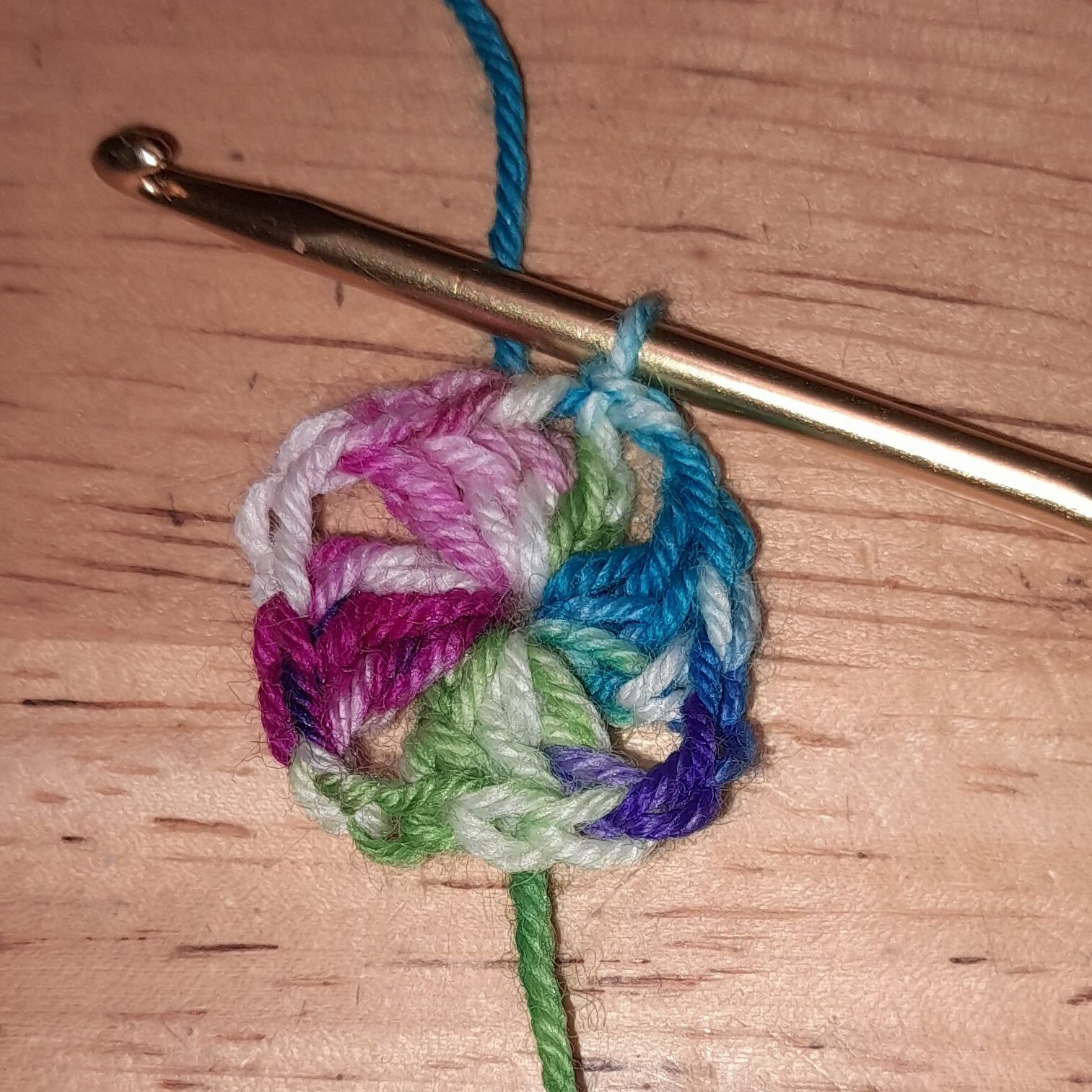 The first round of a crocheted granny square has been worked using a gold crochet hook and a variegated yarn. The square and the hook are laying on a brown table.